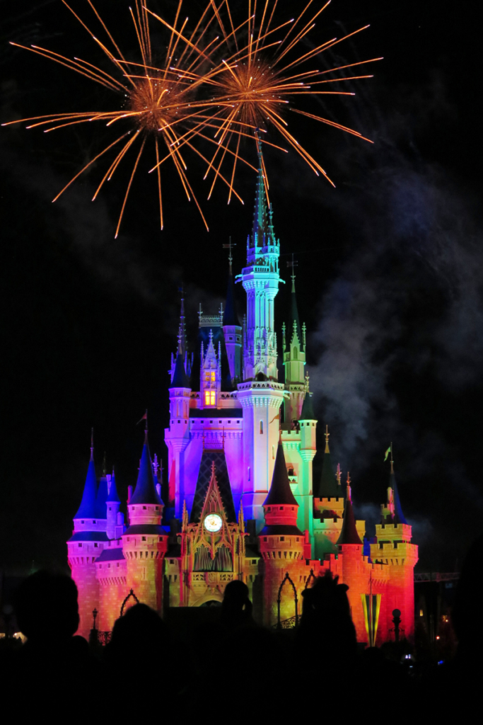 Cinderella's castle at Walt Disney World lit in rainbow colors at night with fireworks in the sky above